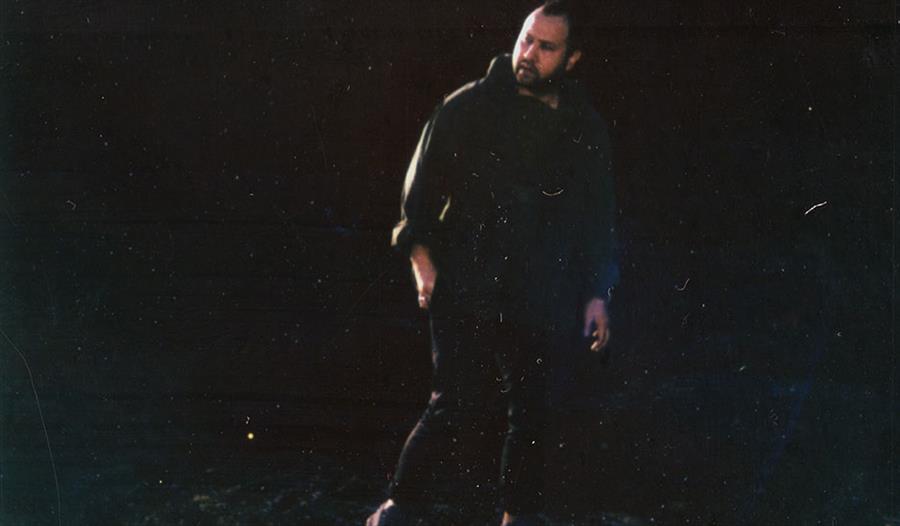 photograph of a white man in a black hoodie, in the shadows against a black background.