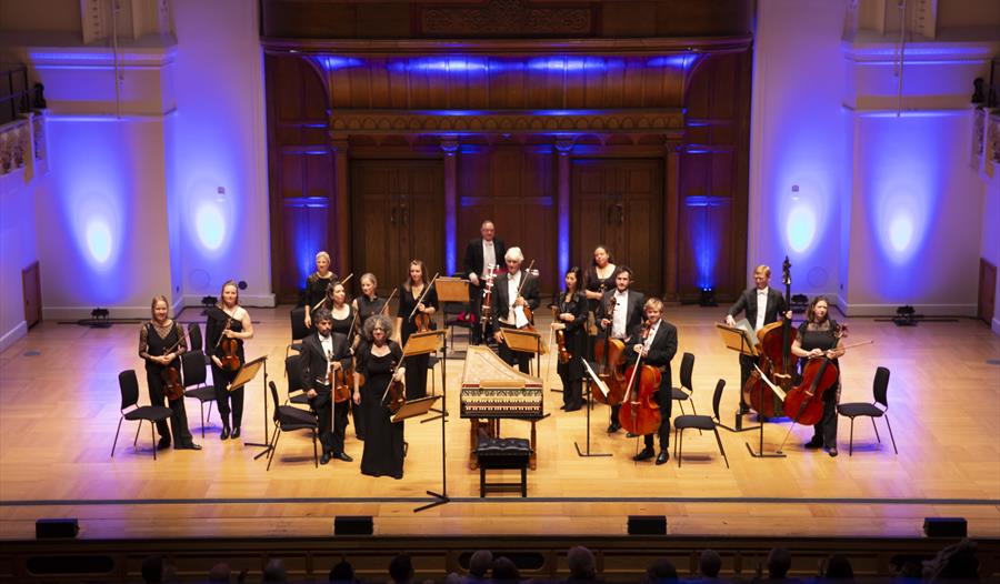 a high up view of an orchestra performing on stage. light wooden flooring, musicians all stand and face toward hidden audience.
