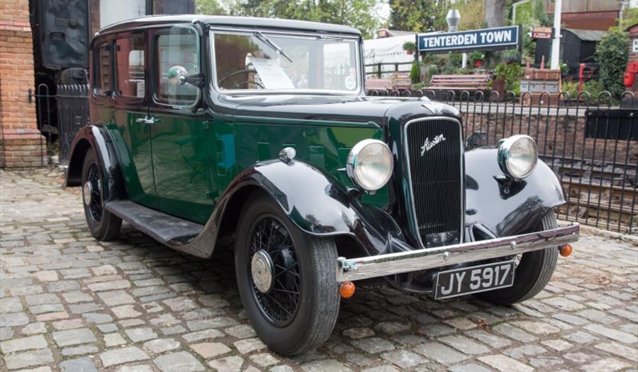 a photograph of a vintage green car parked on brick paving outside a railway station.