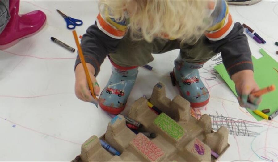 small blond boy in wellies standing on doodled paper with pencil in hand.