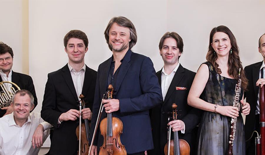 a long group portrait of musicians holding instruments, all white and wearing smart dress. All men except for one woman in silver dress.