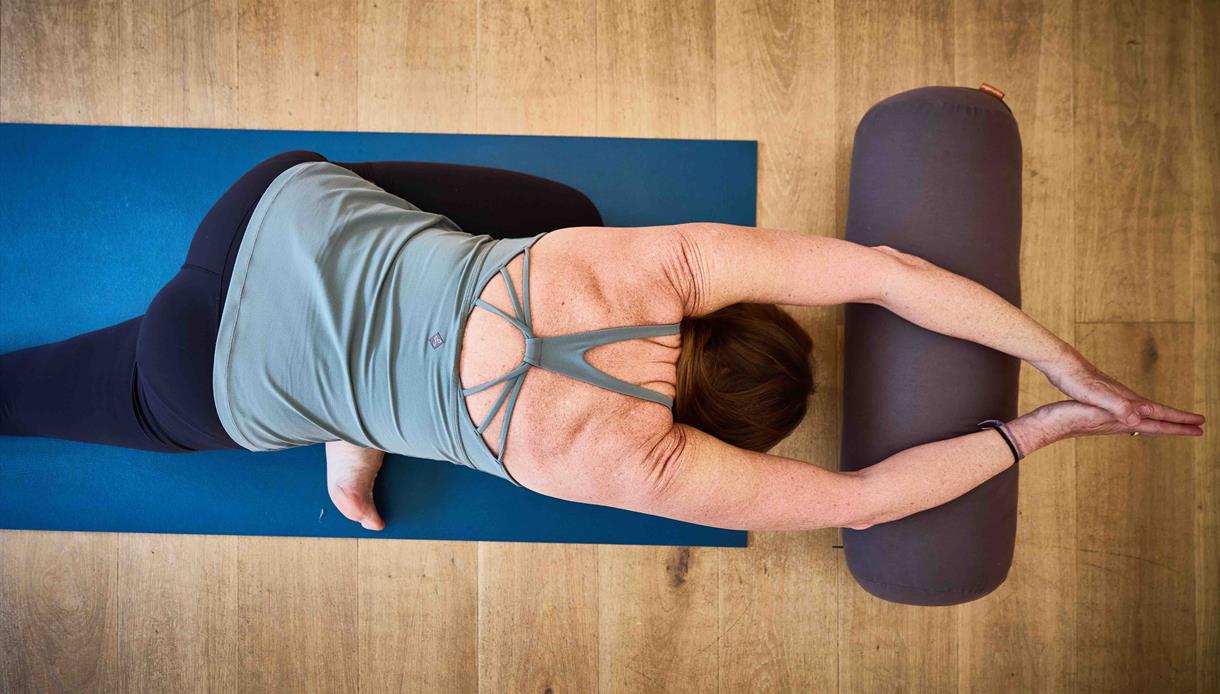 Person practicing a supported yoga stretch on a mat, arms extended overhead and resting on a bolster in a calm indoor setting.