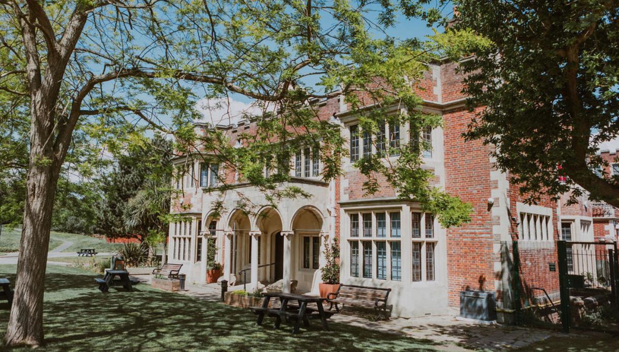 Exterior of Hastings Museum & Art Gallery, a red-brick building set in gardens with trees and picnic benches.