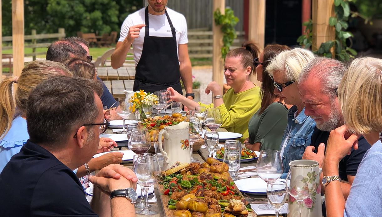 Group of people say at long outdoor picnic table ful lof food. At the head is a man standing, wearing an apron.