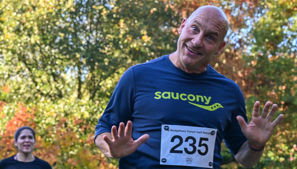 Runner wearing bib raises both hands while crossing a forest trail during a run at Bedgebury National Pinetum.