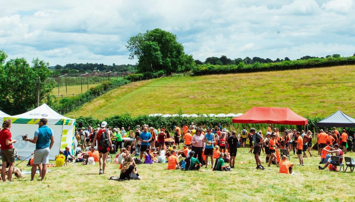 Outdoor event with people gathered under colourful tents on a grassy field with trees and hills in the background.