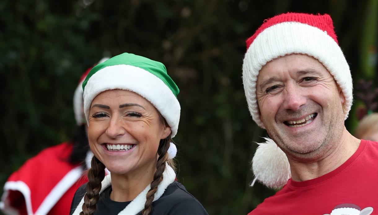 Two people in festive hats smiling at camera outdoors, promoting the Christmas Pudding Dash fun run event.