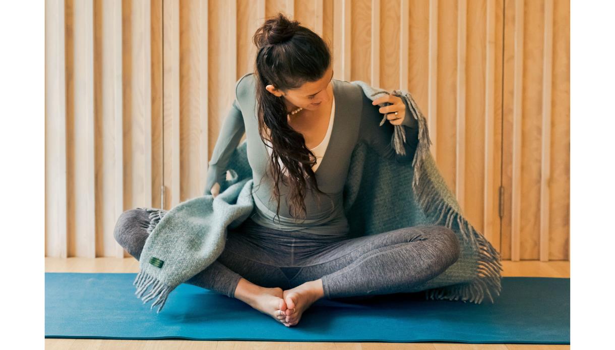Image shows a person sitting on a yoga mat with their legs bent and feet together. They are wearing leggings, a jumper and putting a blanket around th