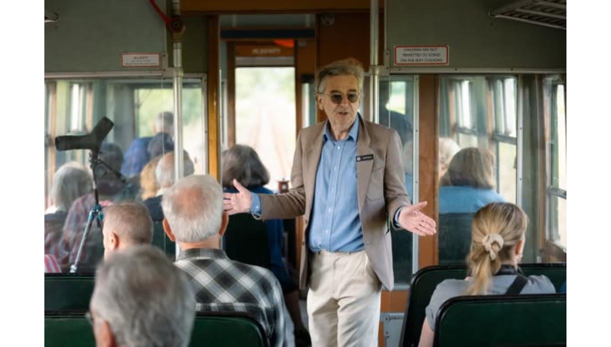 Person standing in the aisle of a vintage train carriage addressing seated passengers.