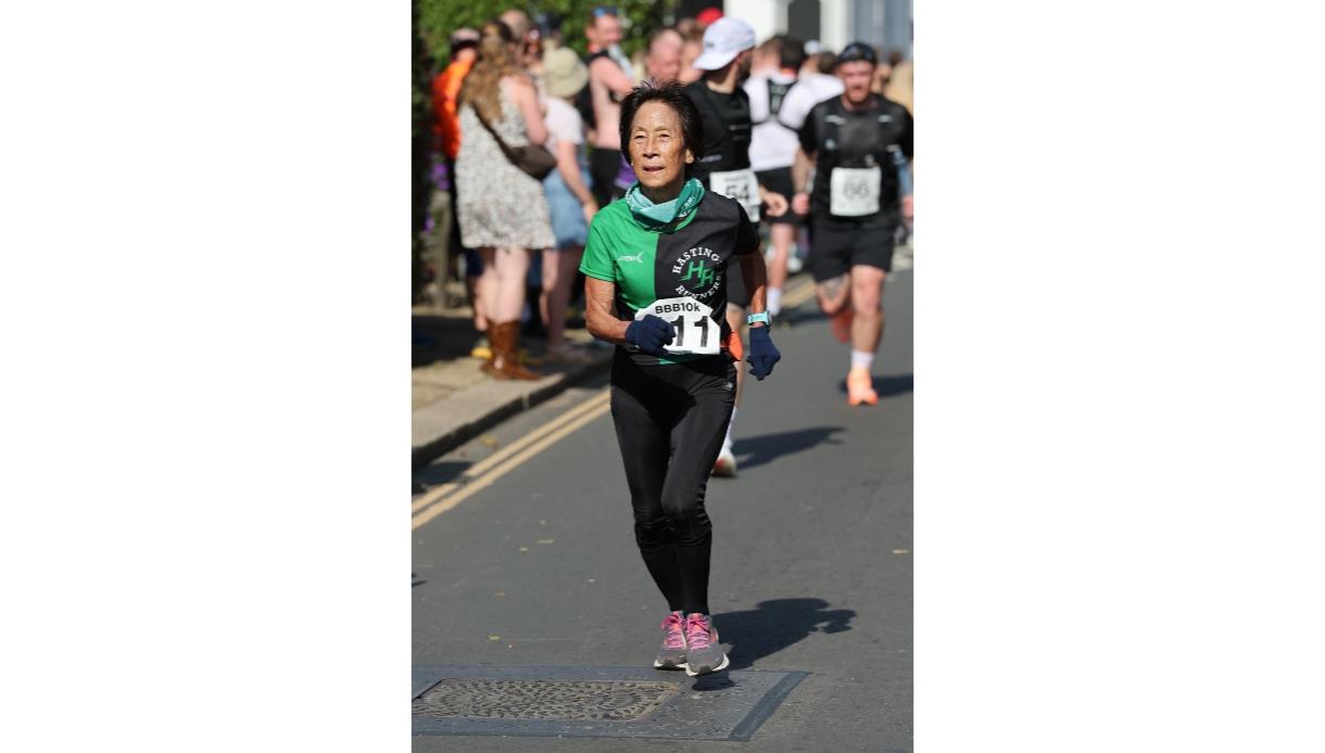 Runner in black and green Hastings Runners shirt with race bib on a sunny street during a 10K event.