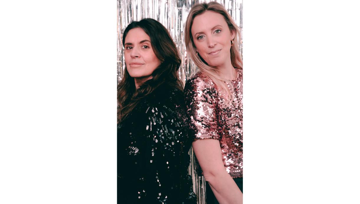 Two people standing side by side in front of a backdrop made of vertical silver metallic streamers. The person on the left is wearing a black sequined