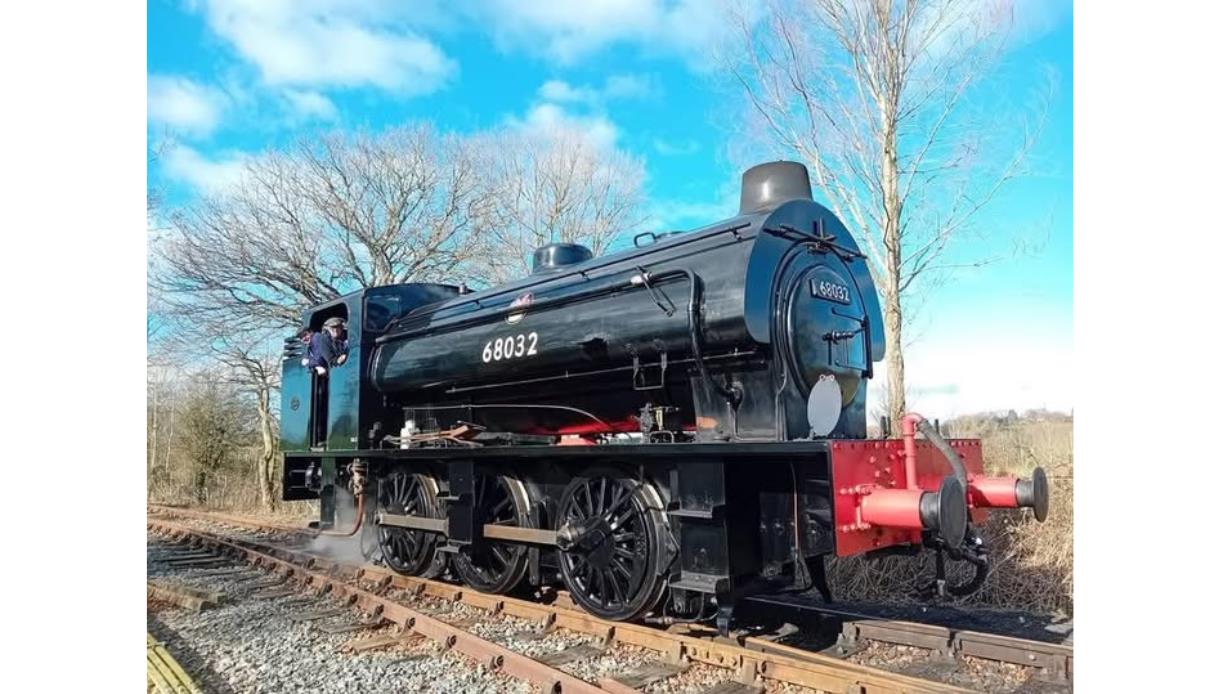 Black steam locomotive on railway tracks with a person leaning from the cab on a bright, clear day.