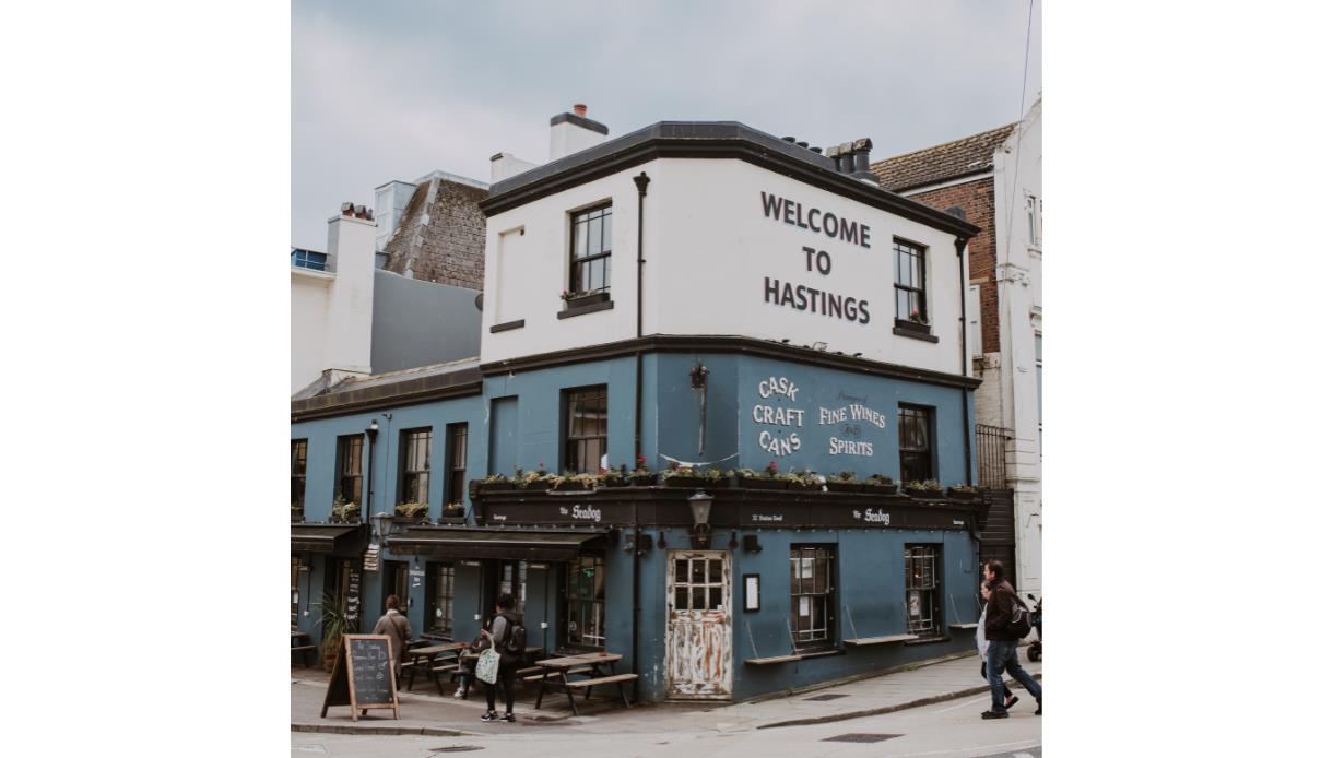 Blue-painted pub with sign reading 'Welcome to Hastings' and outdoor seating on a street corner.