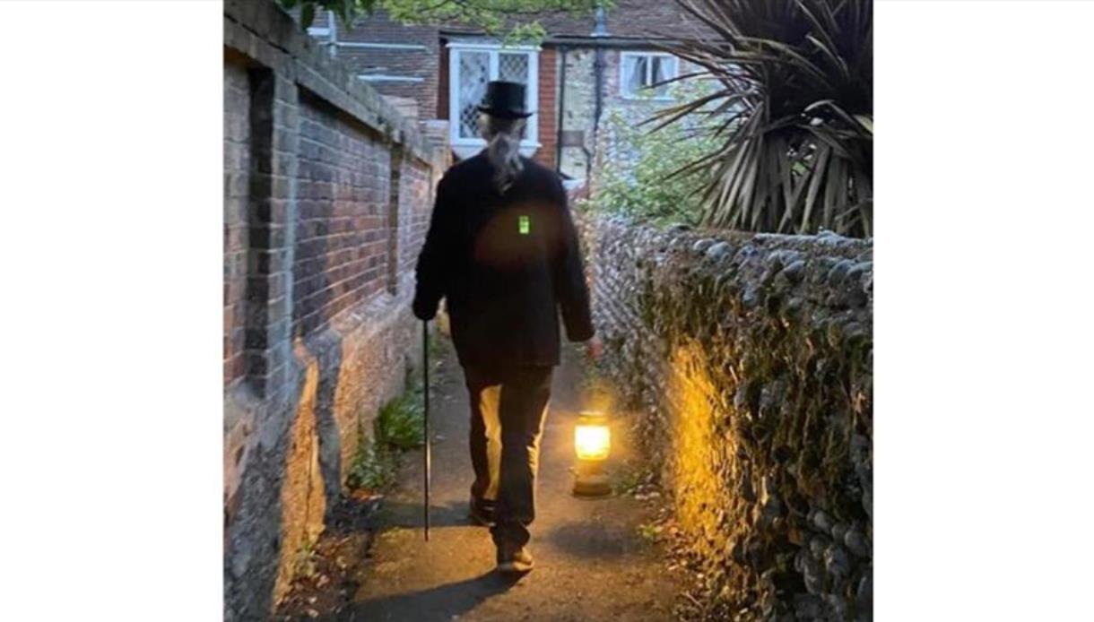 Person in dark coat and top hat walking down a narrow alley with a lantern lighting the path.