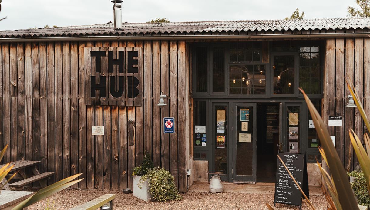 Exterior of The Hub at Quarry Farm near Bodiam, with wooden cladding, entrance doors, signs and an outdoor seating area.