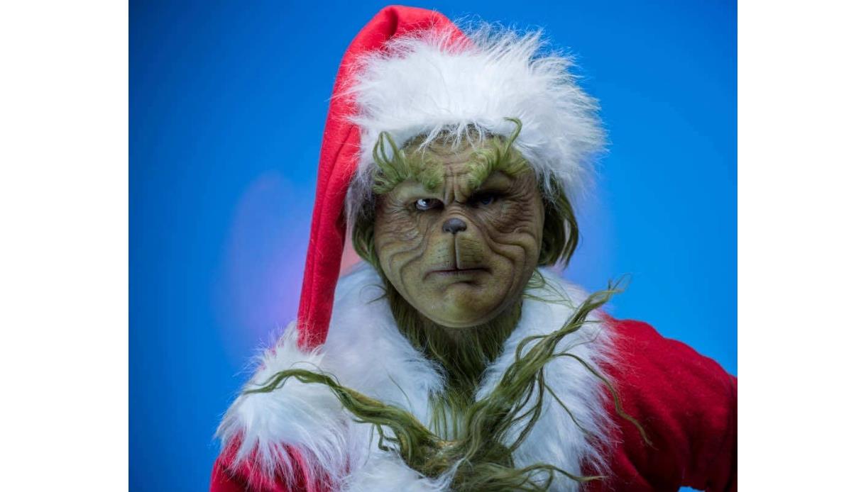 Image shows a picture of 'The Grinch' with green skin wearing a santa outfit which is red with white detail. The background of the image is blue.