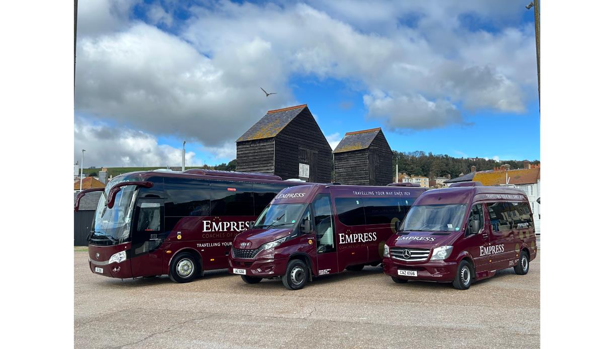 Three maroon Empress Coaches vehicles parked together in front of wooden huts under a cloudy sky