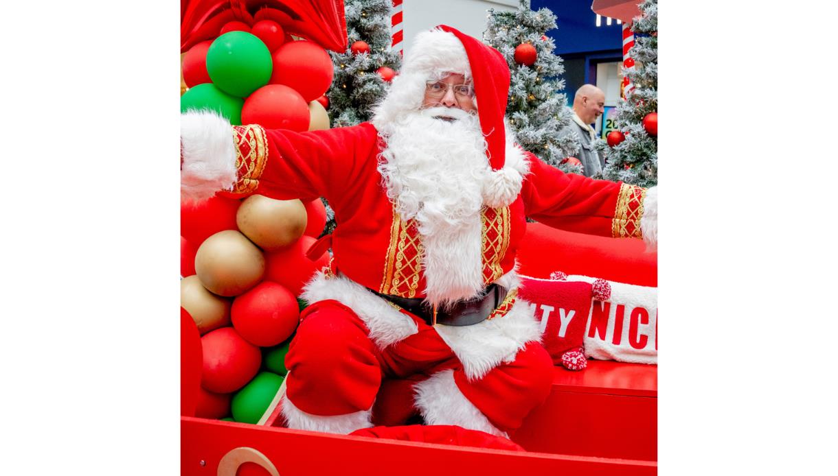 A person dressed as Santa Claus sits on a bright red sleigh surrounded by festive decorations. They wear a traditional red suit with white fur trim, g