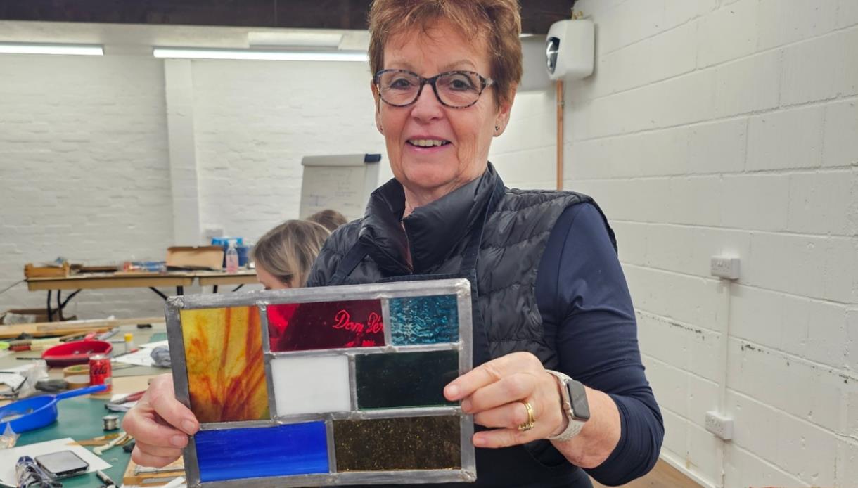 Person holding a handmade stained glass panel in a workshop filled with tools and materials.