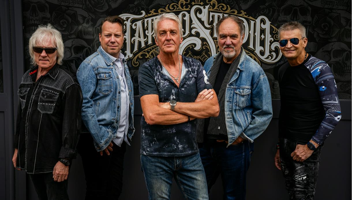 Rock band FM standing together in casual and denim outfits in front of a tattoo‑style backdrop.