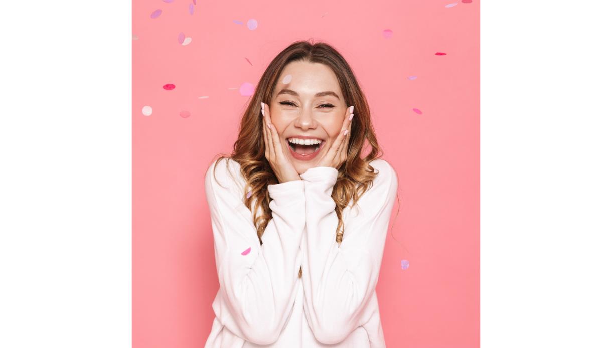 A young woman stands against a bright pink background, smiling widely with her eyes slightly closed and hands pressed to her cheeks in excitement. She