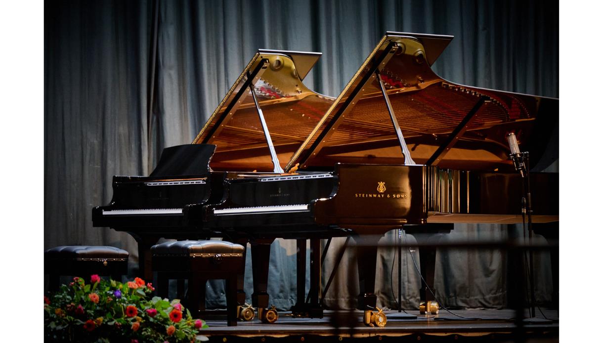 Two grand pianos with open lids on a stage, with stools and flowers in the foreground.