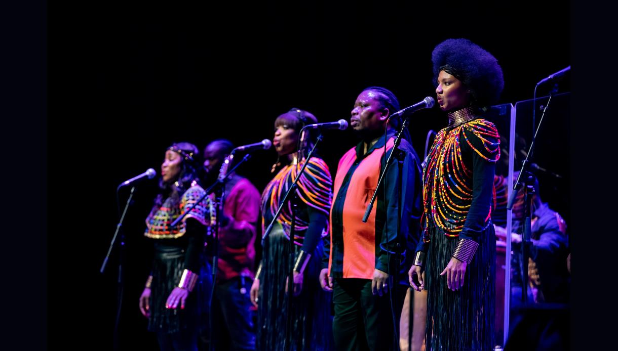 Performers sing into microphones on stage at De La Warr Pavilion, wearing vibrant costumes for Bob Marley reimagined concert.