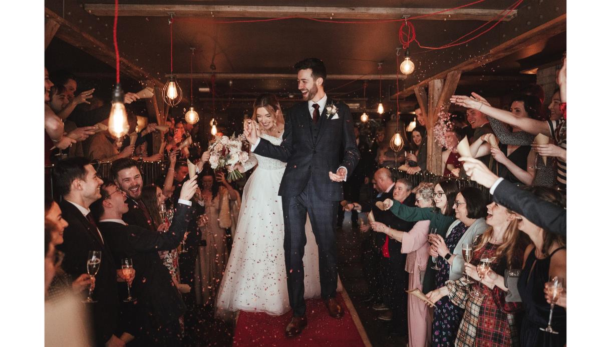 Couple walk through cheering wedding guests at a rustic venue, promoting The Bell’s Quirky Wedding Fair.