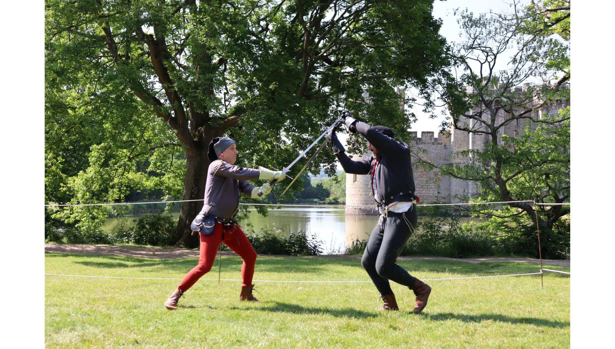 Call to Arms demonstrators stage medieval sword fighting on grass at Bodiam Castle, with the moat and stone walls behind.