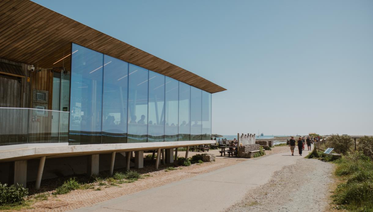Modern glass-fronted Rye Harbour Discovery Centre beside coastal path, with visitors, seating area and sea views.
