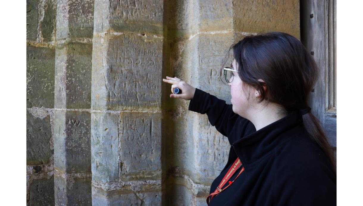 Guide points out carved medieval graffiti on a stone pillar during a tour at Bodiam Castle.