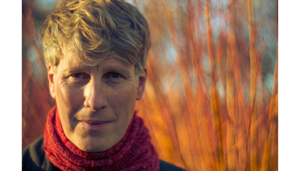 Close-up portrait of Scott Matthews outdoors wearing a red scarf, with short light hair, against a softly blurred background of warm-toned trees.