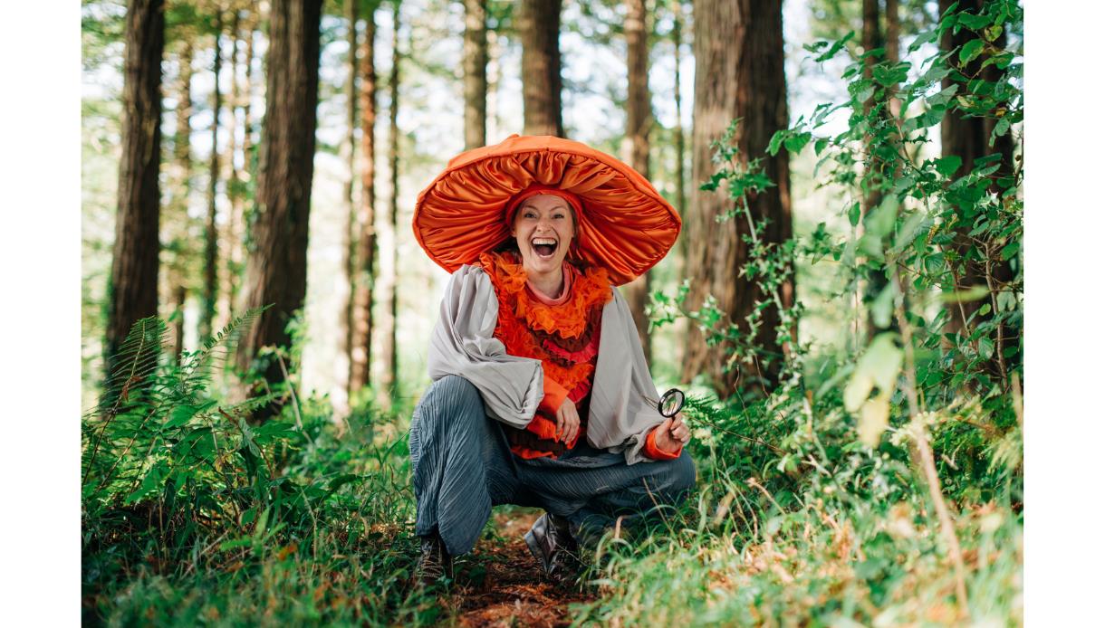 Performer in oversized orange mushroom costume crouching in forest at Bedgebury, holding magnifying glass among trees.