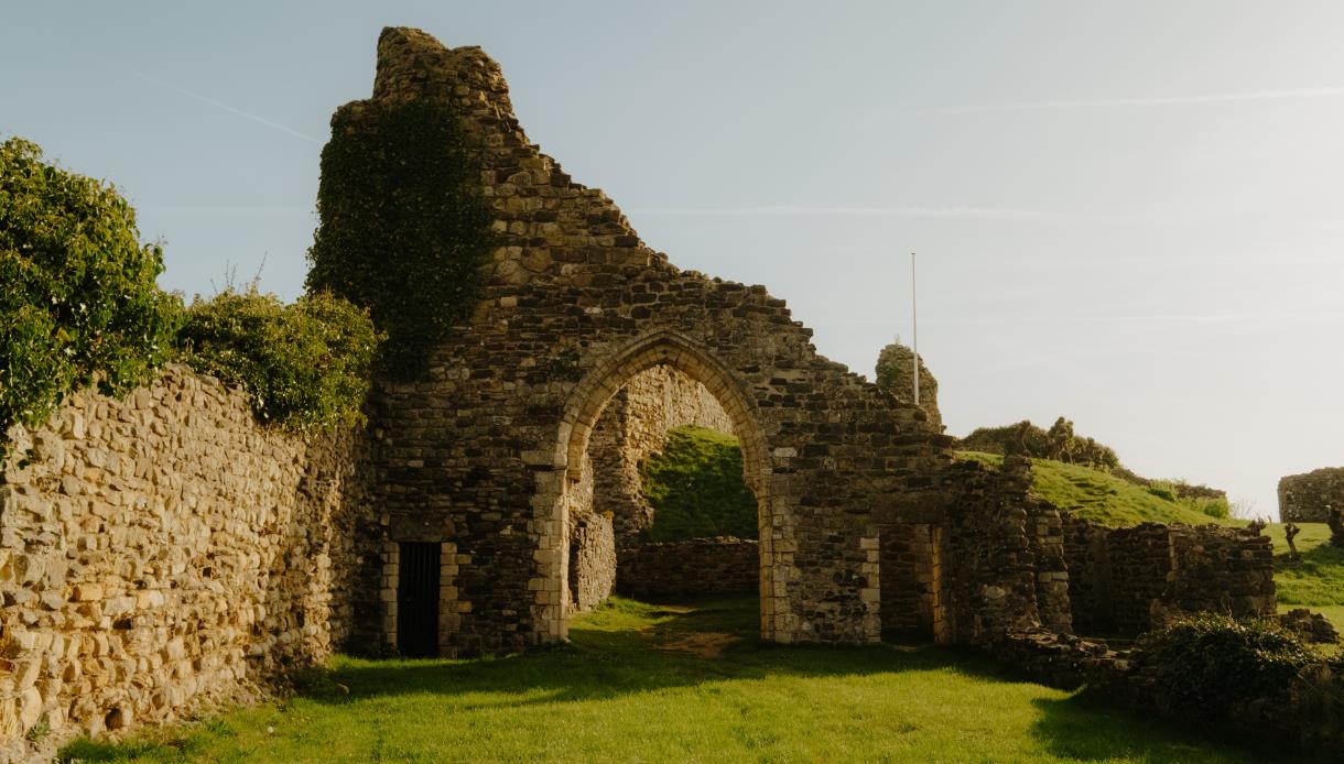Stone archway and ivy-clad ruins of Hastings Castle in warm sunlight, with grassy grounds and hillside beyond.