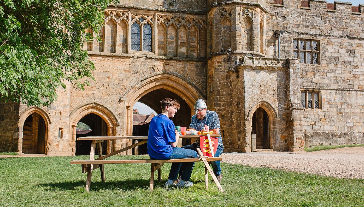 Two visitors sit at a picnic table beside the historic stone arches and medieval architecture of Battle Abbey.