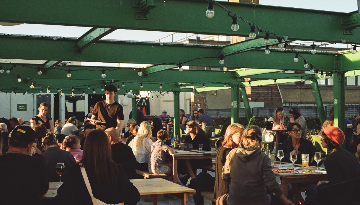 People dining on Bunka Rooftop terrace under green steel beams and string lights in warm evening light.