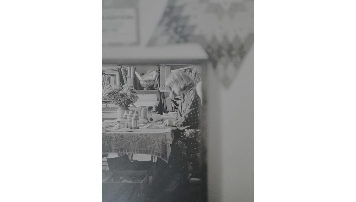 Black-and-white photograph of an artist working at a table in a studio, featured in the Annie Soudain Art Exhibition.