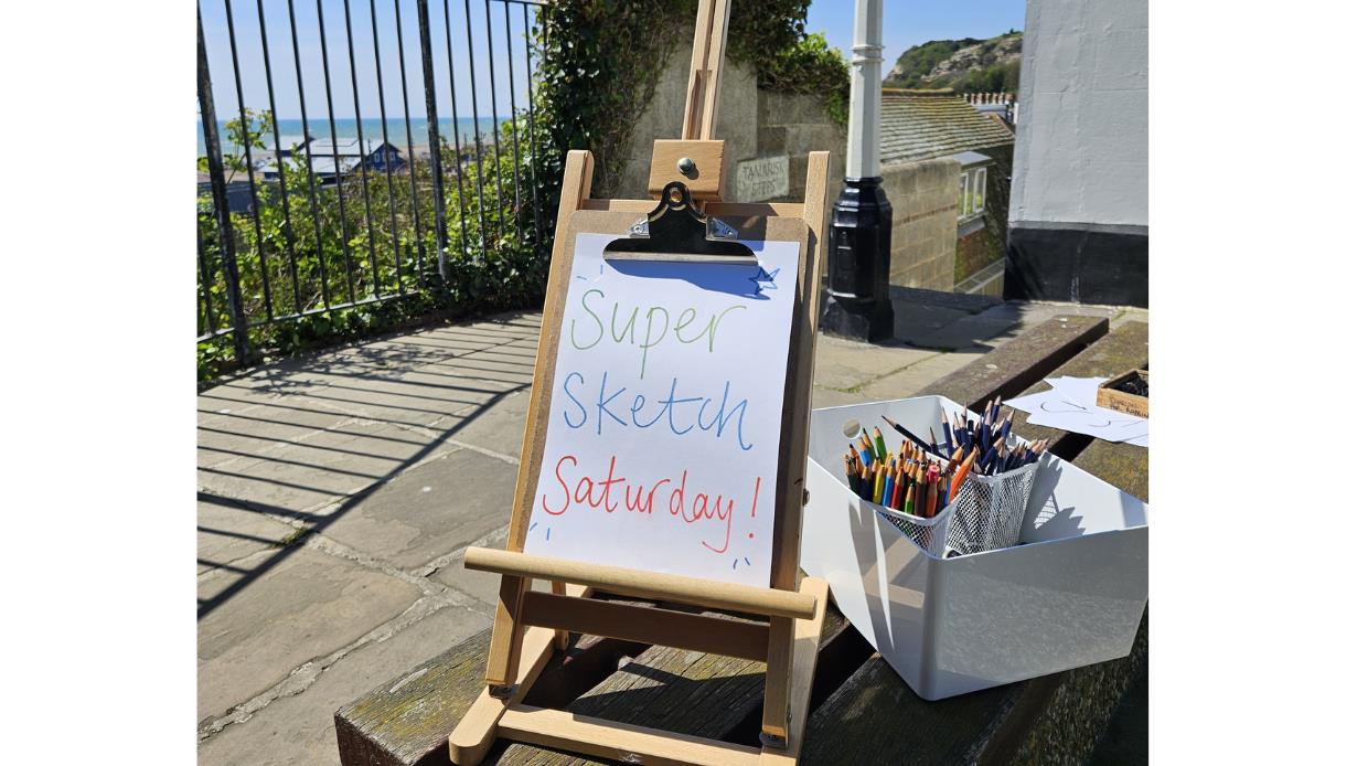 Easel sign reading “Super Sketch Saturday” beside art materials, overlooking the sea at Hastings Contemporary
