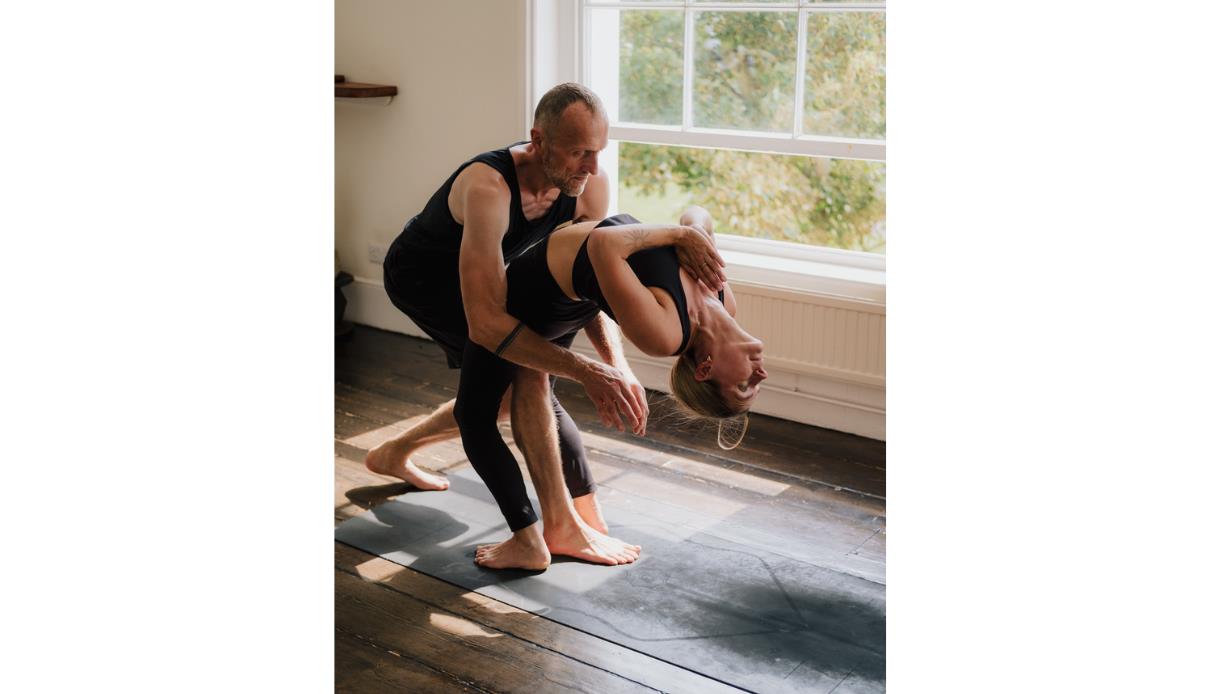 Yoga instructor assisting a participant in a deep backbend during an Ashtanga yoga masterclass in a bright studio.