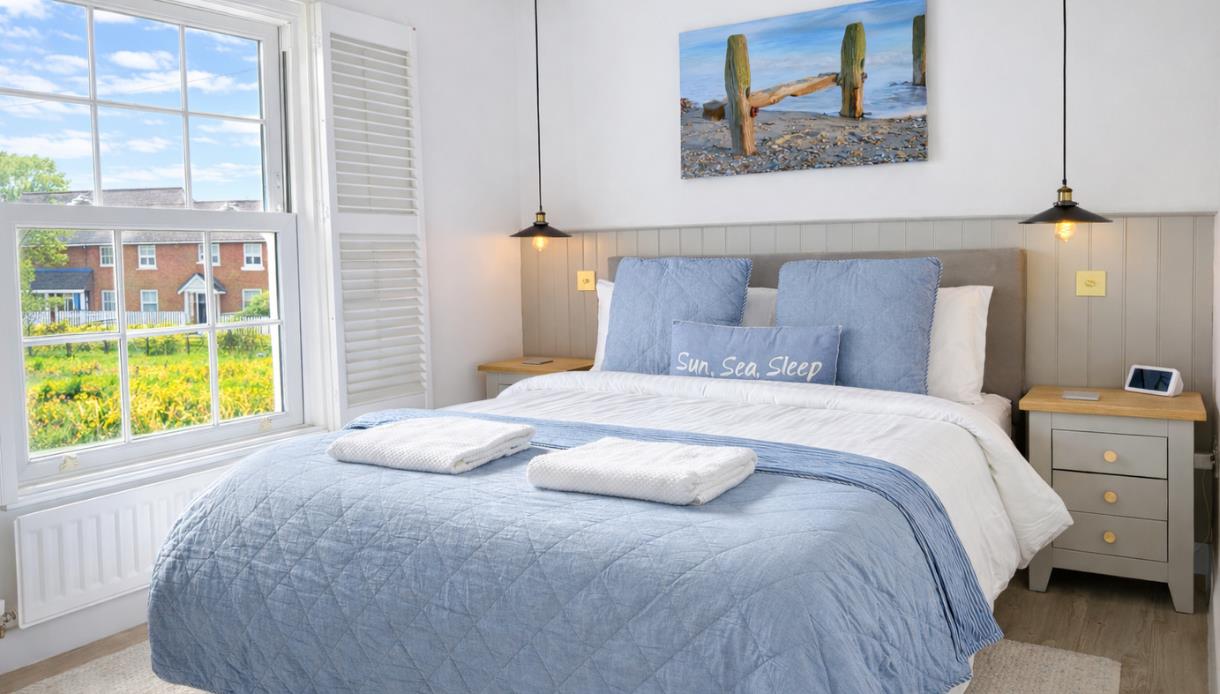 Bright bedroom at Beside The Sea Holidays with a blue bedspread, coastal artwork, and large window overlooking houses