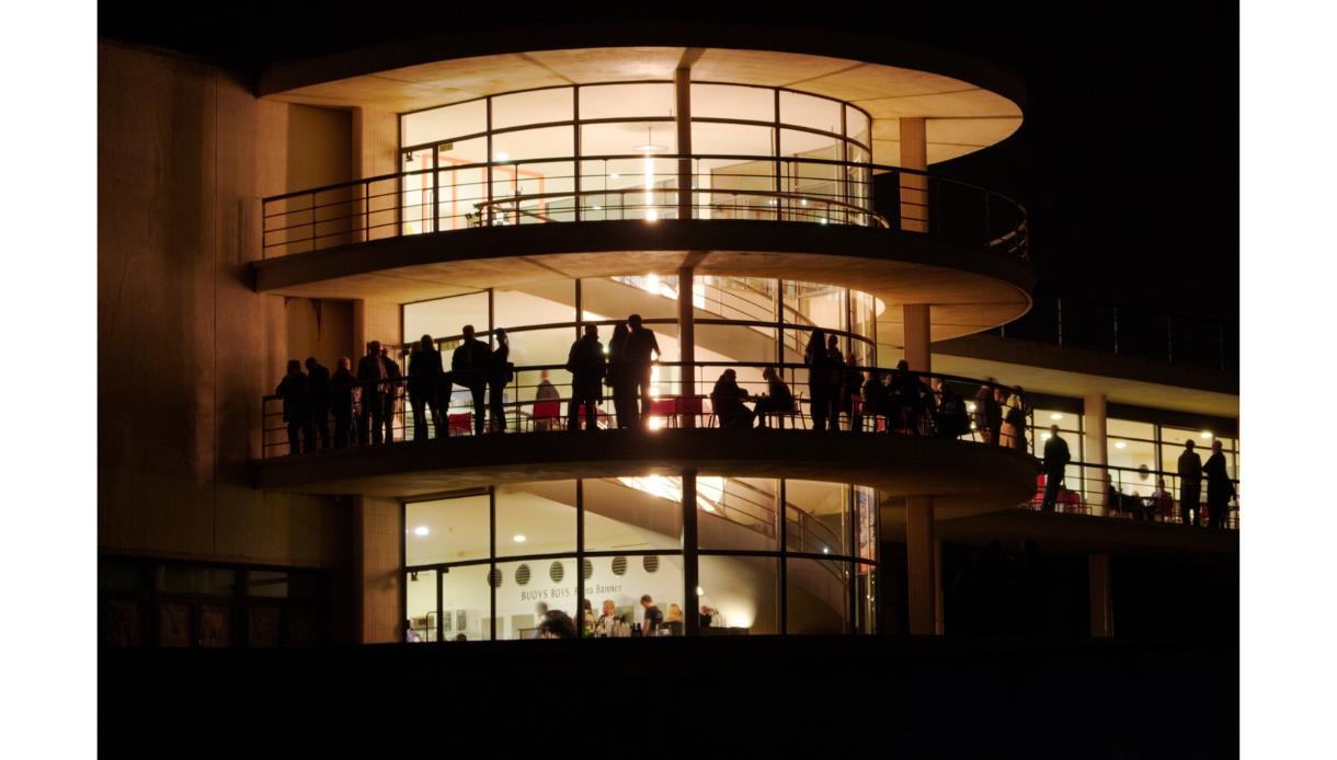 De La Warr Pavilion at night, glowing with interior lights and silhouetted visitors on curved balconies.