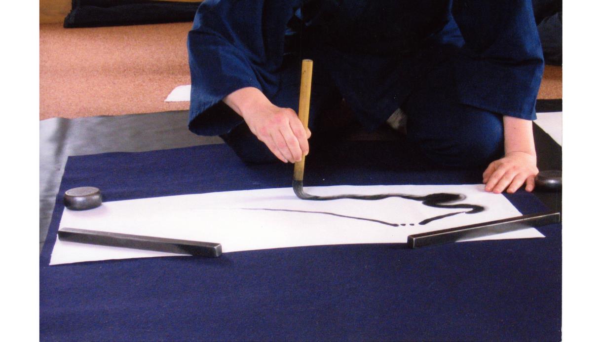 Person practicing Zen brushwork, painting a flowing black ink line on paper during a Summer Zen Brushwork workshop.