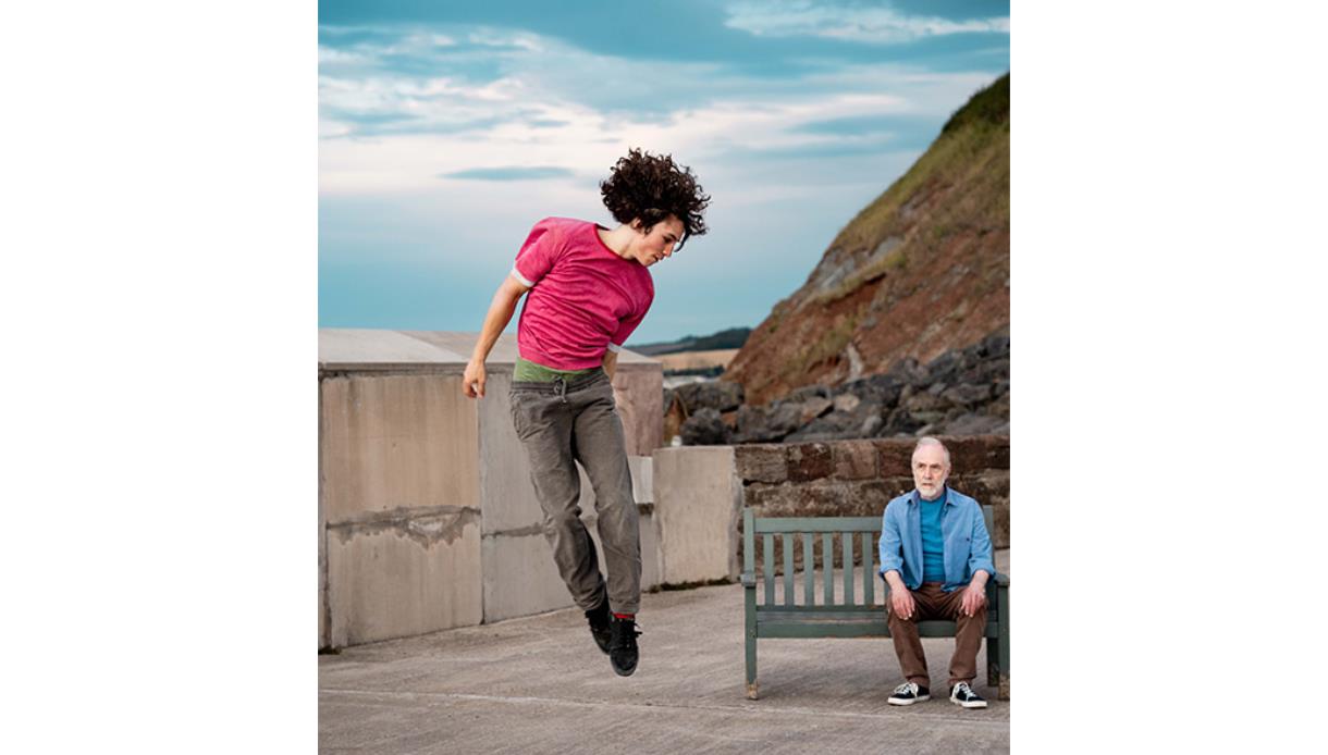 Outdoor dance duet at De La Warr Pavilion, with one performer leaping mid-air beside another seated on a bench by the sea.