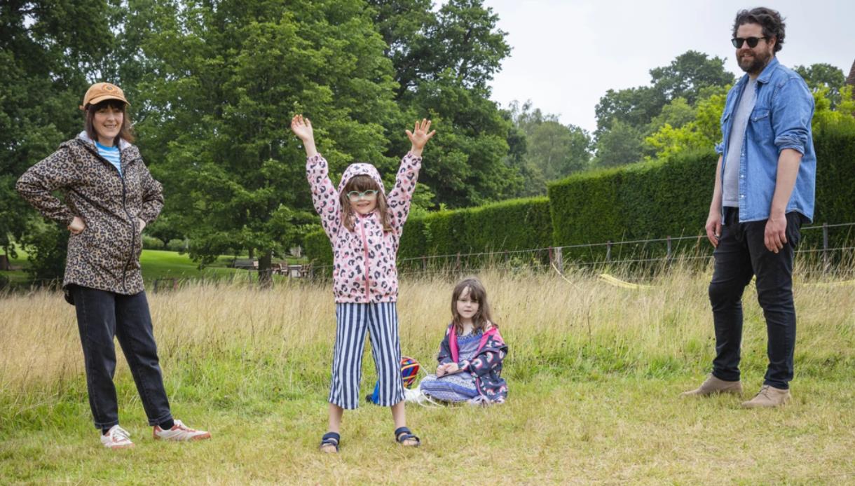 Family outdoors at Bateman's on a grassy field, with two adults and two children spaced apart, trees in the background on a cloudy day.