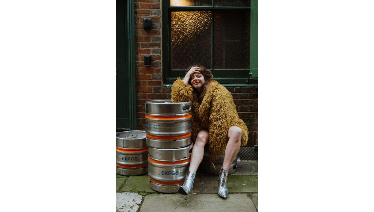 Person in a shaggy coat seated beside metal beer kegs in an urban alleyway with brick walls and green doors.