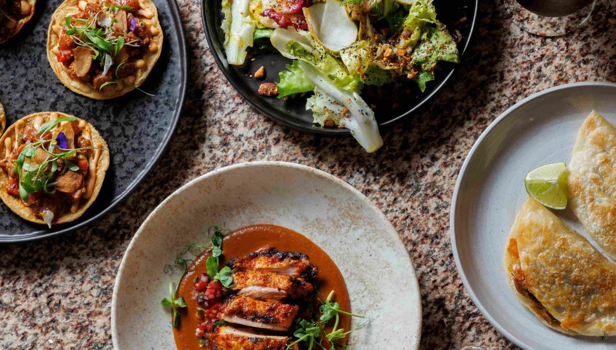 Overhead view of colourful dishes at Crafted, including tacos, grilled chicken, salad and golden empanadas.