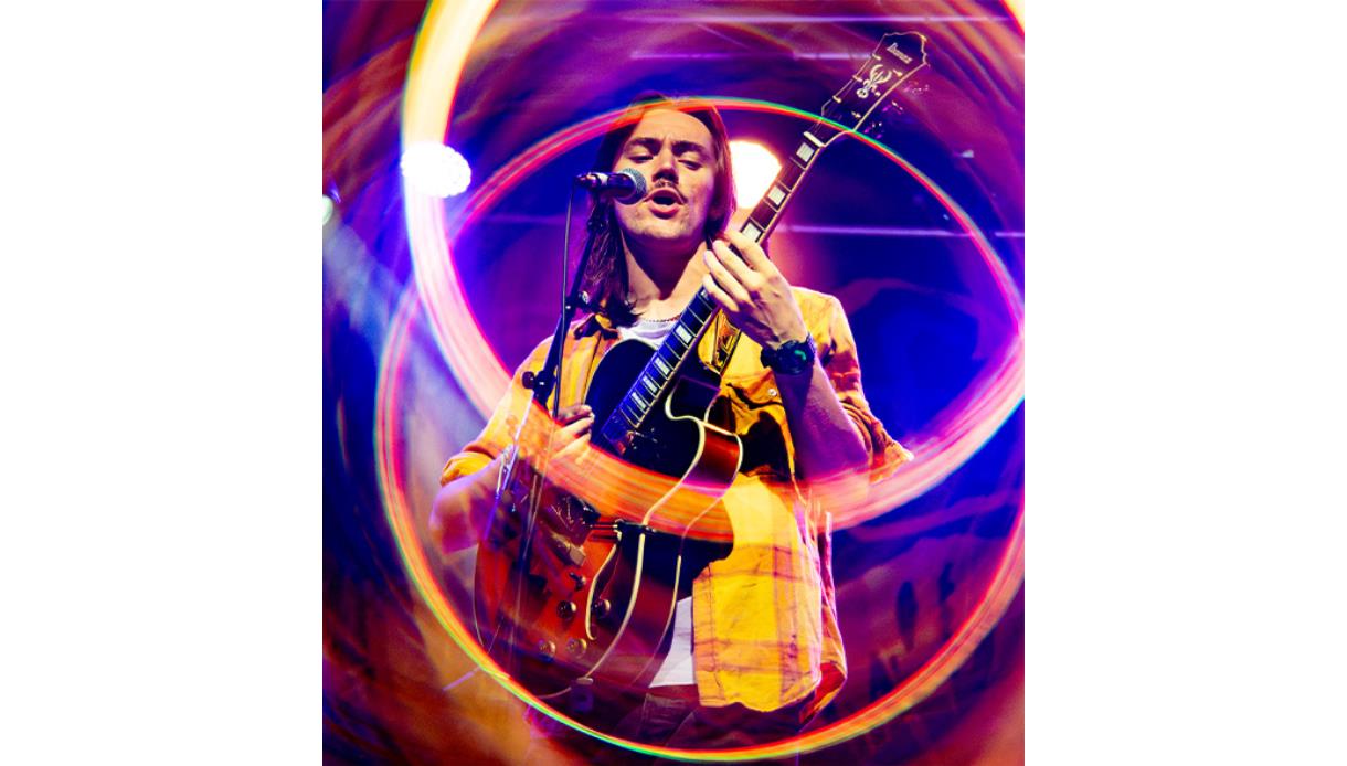 Performer sings while playing electric guitar on stage, surrounded by colourful light rings at Talent Accelerator Summer Sessions, DLWP.
