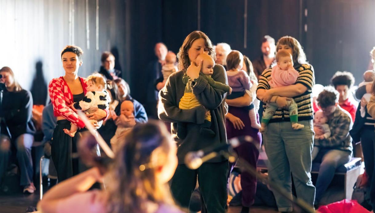 Caregivers holding babies gather around a live performance during A Dance for Us, a free event for babies 0-12 months.