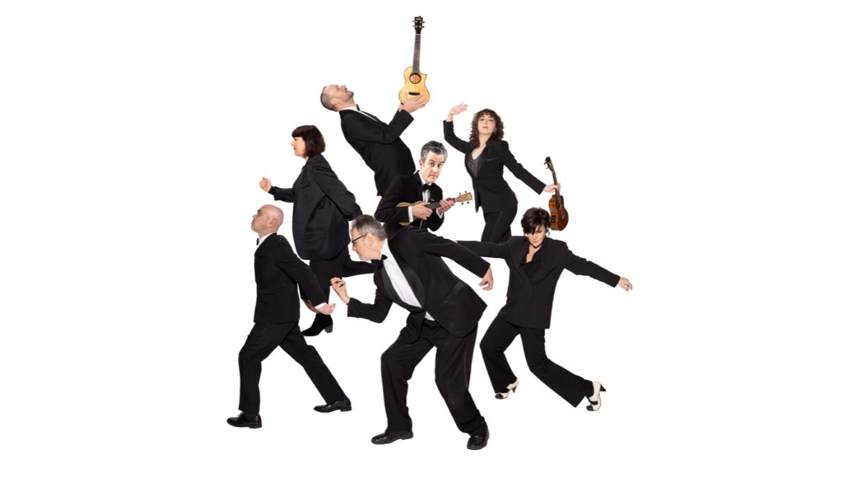 The Ukulele Orchestra of Great Britain in black suits, posed dynamically with ukuleles against a plain white background.