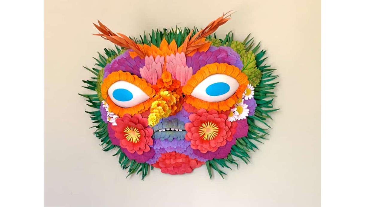 Bright floral mask made from layered paper flowers and leaves, with blue eyes, orange brows, and green leaf edges.